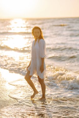 Beyaz kanat çırpan elbiseli genç ve mutlu bir kadın deniz kıyısında yürüyor. Kız sihirli gün doğumuna bakıyor. Yaz zamanı. Seyahat, hafta sonu, rahatlama ve yaşam tarzı konsepti.