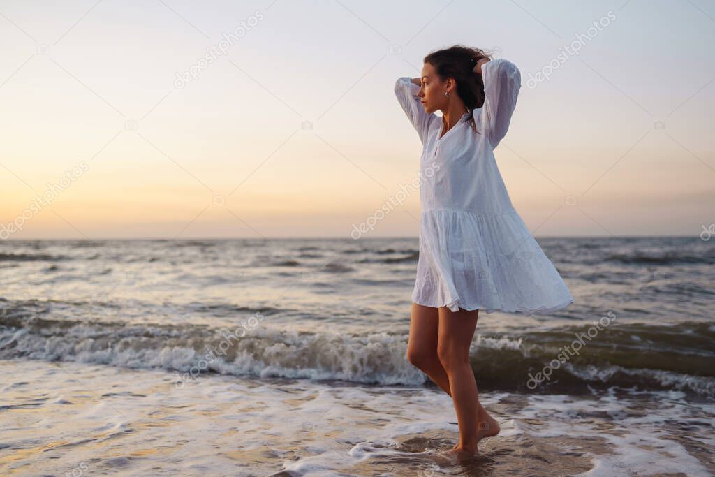 Young happy woman in a white fluttering dress walks along seashore. The girl looks at the magical sunrise. Summer time. Travel, weekend, relax and lifestyle concept.