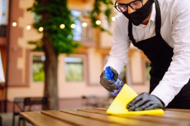 COVID-19 'u önlemek için dezenfekte ediyorum. Koruyucu yüz maskesi takan garson ve eldiven temizleme ve bir sonraki müşteri için restoran masasını dezenfekte etme.