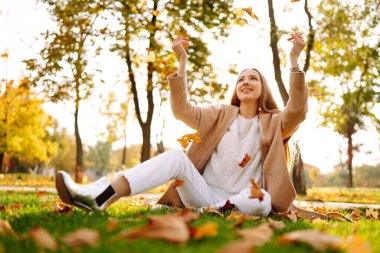 Güneşli bir sonbahar gününde parkta gülümseyen ve yapraklarla eğlenen genç bir kadın. Sonbahar modası. Yaşam tarzı. Rahatla, doğa kavramı..