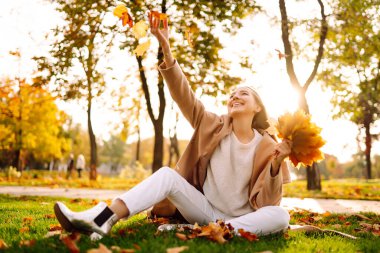 Güneşli bir sonbahar gününde parkta gülümseyen ve yapraklarla eğlenen genç bir kadın. Sonbahar modası. Yaşam tarzı. Rahatla, doğa kavramı..