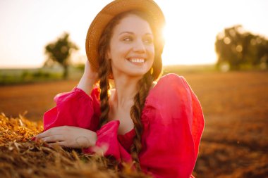  Şapkalı ve elbiseli güzel kıvırcık kadın gün batımında saman balyalarının yanında poz veriyor. Moda konsepti. Doğa, tatil, rahatlama ve yaşam tarzı.