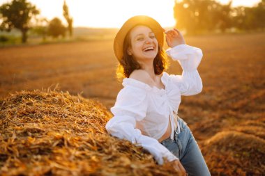  Şapkalı ve elbiseli güzel kıvırcık kadın gün batımında saman balyalarının yanında poz veriyor. Moda konsepti. Doğa, tatil, rahatlama ve yaşam tarzı.