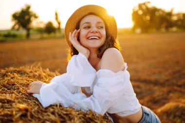  Şapkalı ve elbiseli güzel kıvırcık kadın gün batımında saman balyalarının yanında poz veriyor. Moda konsepti. Doğa, tatil, rahatlama ve yaşam tarzı.