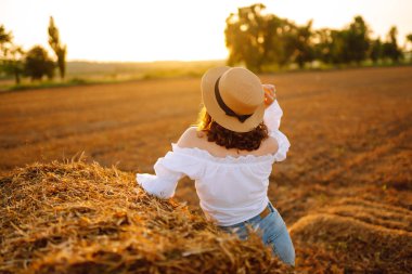  Şapkalı ve elbiseli güzel kıvırcık kadın gün batımında saman balyalarının yanında poz veriyor. Moda konsepti. Doğa, tatil, rahatlama ve yaşam tarzı.