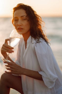 Gün batımında zarif beyaz elbiseler içinde bir bardak şarapla güzel bir manken. Kız sihirli gün doğumuna bakıyor. Yaz zamanı. Seyahat, hafta sonu, rahatlama ve yaşam tarzı konsepti.
