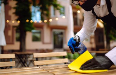 COVID-19 'u önlemek için kafe masalarını dezenfekte ediyorum. Bir kafede çalışırken masaları temizlerken koruyucu maske takan genç bir garson..