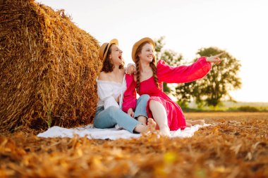 Two smiling young woman resting near haystack. Fashion concept. Nature, vacation, relax and lifestyle.