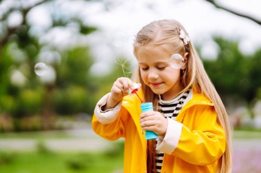 Little girl in a yellow cloak and rubber boots blowing soap bubbles outdoors. Childhood, lifestyle concept.