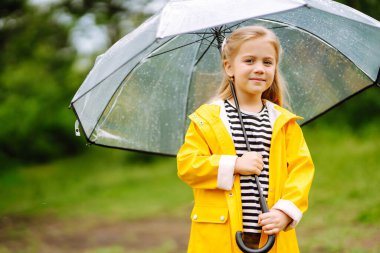 Little girl with transparent umbrella  playing in the rain on sunny autumn day. Beautiful girl  in a yellow cloak and rubber boots. Fall outdoor activity by rainy weather.