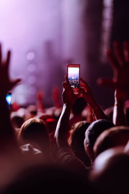 Using a smartphone in a public event,  live music festival.  Holding a mobile phone in hands and shooting photo or video content.  Youth, party, vacation concept.