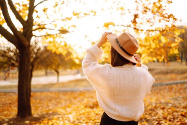 Sonbahar kadını. Beyaz kazaklı ve şapkalı neşeli genç bir kadın gün batımında doğa parkında yürüyor. Moda, tarz kavramı.