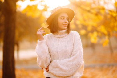 Sonbahar kadını. Beyaz kazaklı ve şapkalı neşeli genç bir kadın gün batımında doğa parkında yürüyor. Moda, tarz kavramı.