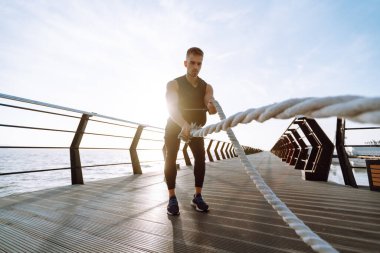 Young man wearing sports clothes is doing exercisesat the beach pier in the morning. Battle ropes. Kettlebell. Sport, Active life.