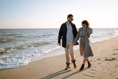 Genç çift, sonbahar güneşli günlerinde kumsalda yürüyüp sarılıyor. Rahatlama, gençlik, aşk, doğayla baş başa yaşam tarzı.