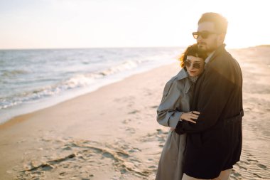 Genç çift, sonbahar güneşli günlerinde kumsalda yürüyüp sarılıyor. Rahatlama, gençlik, aşk, doğayla baş başa yaşam tarzı.