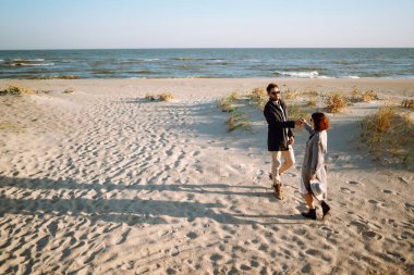 Genç çift, sonbahar güneşli günlerinde kumsalda yürüyüp sarılıyor. Rahatlama, gençlik, aşk, doğayla baş başa yaşam tarzı.