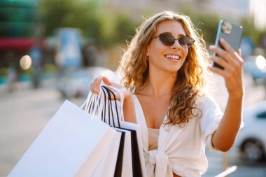 Alışveriş torbalı güzel bir kadın selfie çekiyor. Alışveriş yaptıktan sonra şehirde dolaşan genç bir kadın. Kara Cuma, satış, tüketici, yaşam tarzı konsepti.