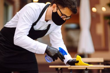 COVID-19 'u önlemek için dezenfekte ediyorum. Garson, restoranda koruyucu maske ve eldivenlerle dezenfektanla masayı temizliyor..