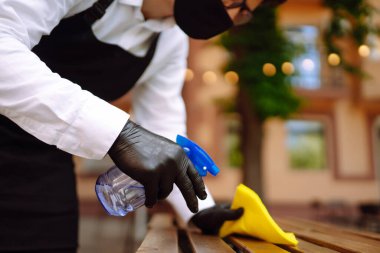 COVID-19 'u önlemek için dezenfekte ediyorum. Garson, restoranda koruyucu maske ve eldivenlerle dezenfektanla masayı temizliyor..