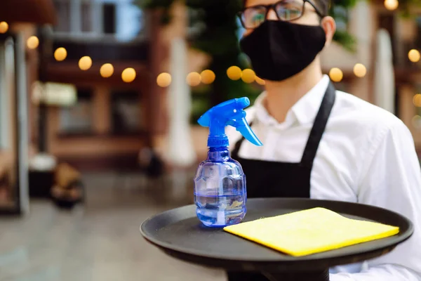 Covid 19を防ぐために消毒します 医療用マスクと手袋を着用レストランで消毒スプレーでテーブルを掃除するウェイター — ストック写真