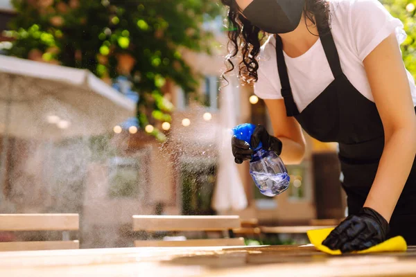 COVID-19 'u önlemek için dezenfekte ediyorum. Garson, restoranda koruyucu maske ve eldivenlerle dezenfektanla masayı temizliyor..