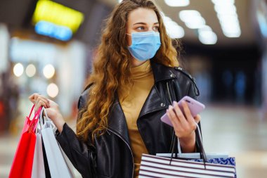 Telefonunu kullanarak alışveriş torbaları taşıyan koruyucu tıbbi maskeli şık bir kadın. Sonbahar alışverişi. Moda, tarz, Kara Cuma, satış, tüketim konsepti.