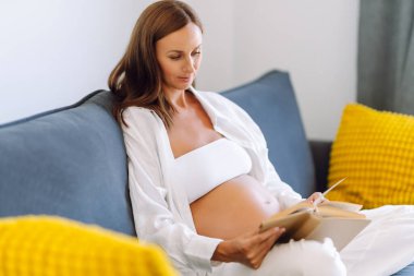 A beautiful woman expecting baby is reading a book while lying on sofa in cozy living room. Pregnant woman enjoys a book and relaxes indoors. The concept is pregnancy, relaxation, and comfort.