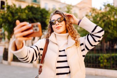 A happy woman in stylish clothes takes a selfie on phone in beautiful city square. A beautiful tourist enjoys sunset and blogs while strolling through city streets. Concepts of tourism and blogging.