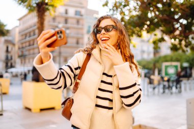 A happy woman in stylish clothes takes a selfie on phone in beautiful city square. A beautiful tourist enjoys sunset and blogs while strolling through city streets. Concepts of tourism and blogging.