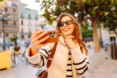A happy woman in stylish clothes takes a selfie on phone in beautiful city square. A beautiful tourist enjoys sunset and blogs while strolling through city streets. Concepts of tourism and blogging.