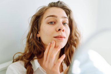 A close-up of a young woman's face looks into a small mirror in cozy room. The beautiful woman touches face with hands, enjoying her reflection. Concept of beauty and relaxation. Cosmetology.