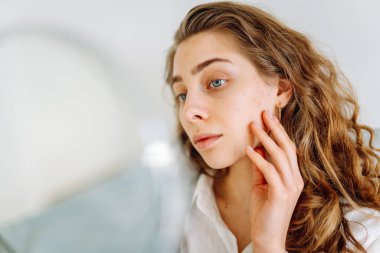 A close-up of a young woman's face looks into a small mirror in cozy room. The beautiful woman touches face with hands, enjoying her reflection. Concept of beauty and relaxation. Cosmetology.