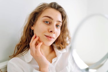 A close-up of a young woman's face looks into a small mirror in cozy room. The beautiful woman touches face with hands, enjoying her reflection. Concept of beauty and relaxation. Cosmetology.