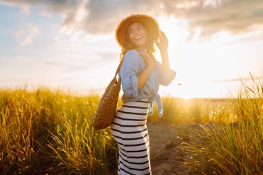 A young woman in a straw hat strolls along the beach on a sunny day. A beautiful woman relaxes and enjoys the sunset. Concept of relaxation and enjoyment. Lifestyle.