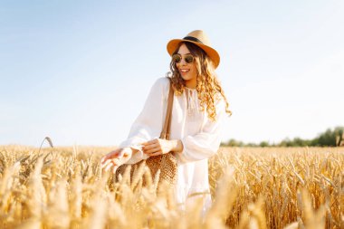 A beautiful woman in a white dress and hat enjoys a walk through a wheat field. A joyful woman is having fun in a farm field. Concept of tranquility, style and nature.