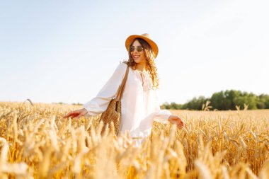 A beautiful woman in a white dress and hat enjoys a walk through a wheat field. A joyful woman is having fun in a farm field. Concept of tranquility, style and nature.