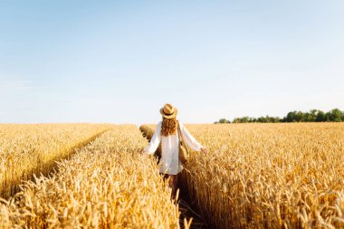A beautiful woman in a white dress and hat enjoys a walk through a wheat field. A joyful woman is having fun in a farm field. Concept of tranquility, style and nature.