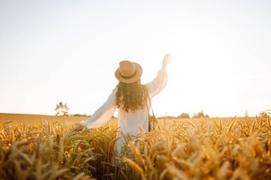 A beautiful woman in a white dress and hat enjoys a walk through a wheat field. A joyful woman is having fun in a farm field. Concept of tranquility, style and nature.