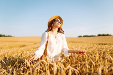 A beautiful woman in a white dress and hat enjoys a walk through a wheat field. A joyful woman is having fun in a farm field. Concept of tranquility, style and nature.