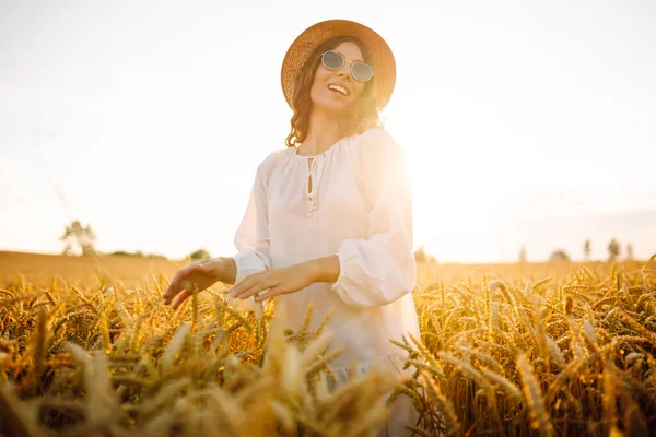 A beautiful woman in a white dress and hat enjoys a walk through a wheat field. A joyful woman is having fun in a farm field. Concept of tranquility, style and nature.