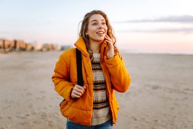 A cheerful traveler in a bright jacket stands on the beach at sunrise. A rear view of the beautiful woman enjoys the sunset and feels the freedom of the outdoors. Concept of adventure and freedom.