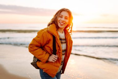 A cheerful traveler in a bright jacket stands on the beach at sunrise. A rear view of the beautiful woman enjoys the sunset and feels the freedom of the outdoors. Concept of adventure and freedom.