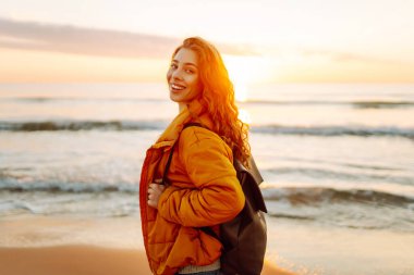 A cheerful traveler in a bright jacket stands on the beach at sunrise. A rear view of the beautiful woman enjoys the sunset and feels the freedom of the outdoors. Concept of adventure and freedom.