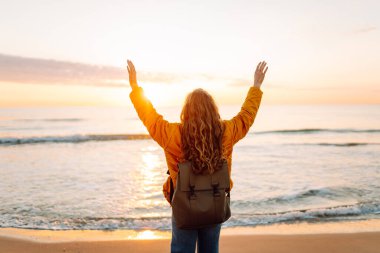 A cheerful traveler in a bright jacket stands on the beach at sunrise. A rear view of the beautiful woman enjoys the sunset and feels the freedom of the outdoors. Concept of adventure and freedom.