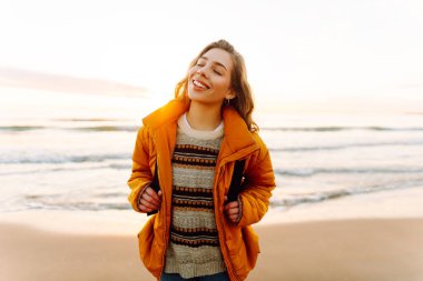 A cheerful traveler in a bright jacket stands on the beach at sunrise. A rear view of the beautiful woman enjoys the sunset and feels the freedom of the outdoors. Concept of adventure and freedom.