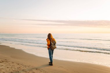 A cheerful traveler in a bright jacket stands on the beach at sunrise. A rear view of the beautiful woman enjoys the sunset and feels the freedom of the outdoors. Concept of adventure and freedom.