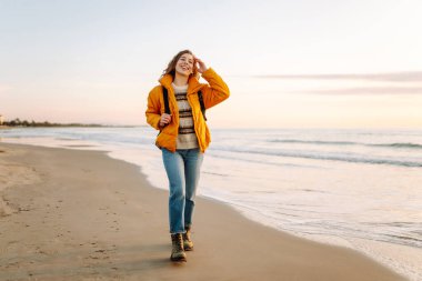 A cheerful traveler in a bright jacket stands on the beach at sunrise. A rear view of the beautiful woman enjoys the sunset and feels the freedom of the outdoors. Concept of adventure and freedom.