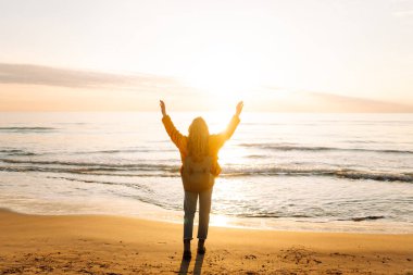 A cheerful traveler in a bright jacket stands on the beach at sunrise. A rear view of the beautiful woman enjoys the sunset and feels the freedom of the outdoors. Concept of adventure and freedom.