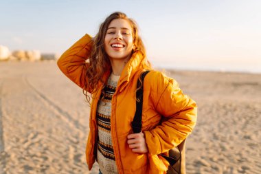 A cheerful traveler in a bright jacket stands on the beach at sunrise. A rear view of the beautiful woman enjoys the sunset and feels the freedom of the outdoors. Concept of adventure and freedom.